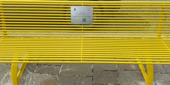 A bright yellow metal bench on a paved square, a small engraved plaque fixed to the middle of its back.